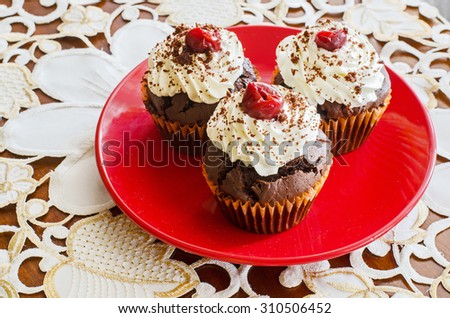 side view of three chocolate muffins with whipped cream and cherry on top on a red plate on a white decorated table cloth