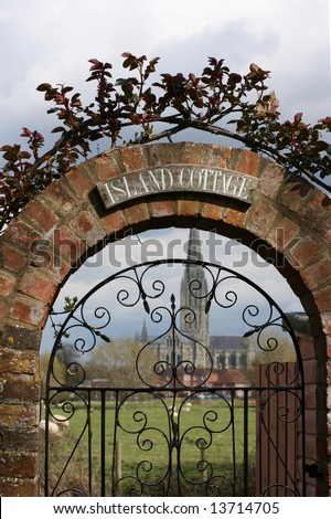 A picturesque cottage gate with arched brick work above covered with a climbing rose, with a view of Salisbury Cathedral visible through the wrought iron gate.