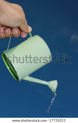 A small hand held watering can being held aloft against a blue sky backdrop. Water emerging from spout of watering can. Portrait format.