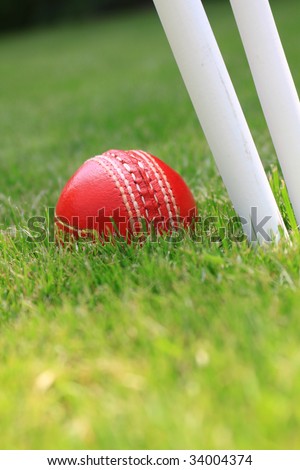 A red leather cricket ball lying in green grass behind the base of three white cricket stumps. Set on a portrait format.