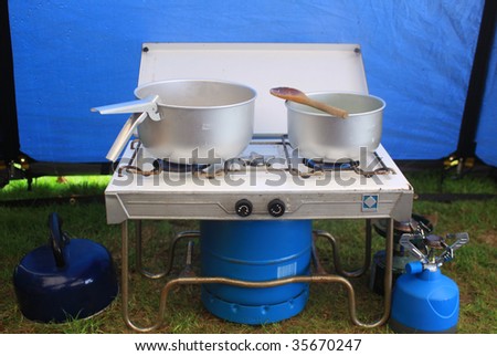 A two ringed gas camping stove. Set inside a blue canvas tent. Two tin pots are on the stove .