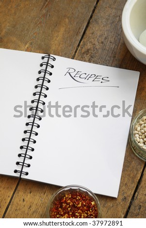 A blank wire spiral bound recipe book with the title \'recipe\' hand written at the top of the page. Set on a wooden kitchen table top. Glass dishes of dried chillies and dried white beans also visible.