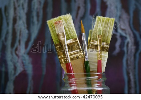 A set of artists brushes in a glass jar, set in front of an abstract oil on canvas painting (by self) in soft focus. Shallow depth of field. Set on a landscape format.