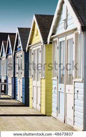 Front view of a row of wooden beach huts with wooden terraces located in Christchurch Hampshire UK. Huts painted in pale blue and yellow colours.