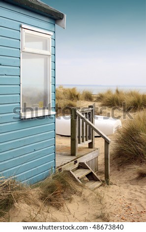Viw of the side of a blue wooden beach hut with wooden terrace, looking towards the coast/beach. A white upturned boat rests in front of the hut amongst the sand and reed bushes.