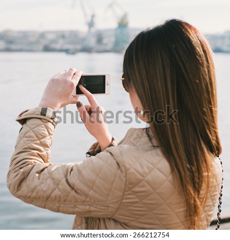 Fashion  woman taking photo with cellphone on the coast on spring. Happy girl on vacation taking picture on sea background. Photo with instagram style filters