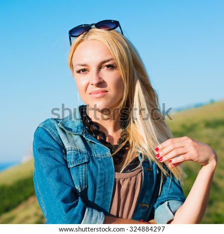 portrait of the attractive, slender, beautiful young Caucasian  blonde girl in a jeans jacket. Smiling girl enjoys fine warm summer weather highly in mountains against the sea
