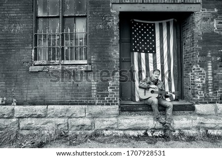 Man looking at camera sitting on brick factory loading dock in jeans with acoustic guitar and American flag, freedom, country, usa, independence, playing guitar outside, singing, portrait, music Сток-фото © 