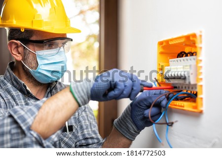 Electrician worker at work on an electrical panel protected by helmet, safety goggles and gloves; wear the surgical mask to prevent the spread of Coronavirus. Construction industry-Covid 19 Prevention