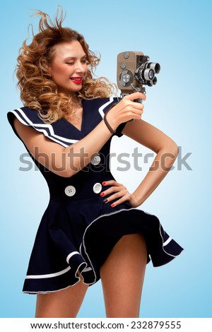 Retro photo of a glamorous pin-up sailor girl with an old vintage cinema 8 mm camera, standing in the wind, shooting a movie on blue background.