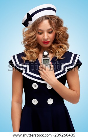 Retro photo of a glamorous pin-up sailor girl holding an old vintage handheld digital flash meter and measuring the correct exposure for the photograph on blue background.