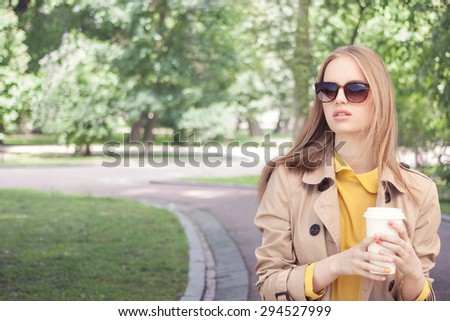 Young fashionable woman taking a coffee break after shopping, walking with a coffee-to-go in her hands against green city park background.