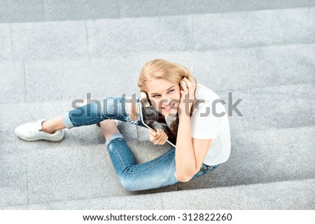 Top view of a happy young woman, listening to the music in vintage music headphones, surfing internet on a tablet pc and sitting on stairs against urban city background.
