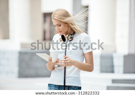 Happy young woman with vintage music headphones around her neck, holding a take away coffee cup and surfing internet on a tablet pc against urban city background.