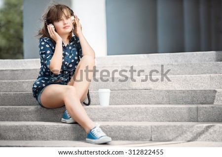 Happy young woman with vintage music headphones, listening to the music and sitting on stairs with a take away coffee cup against urban city background.