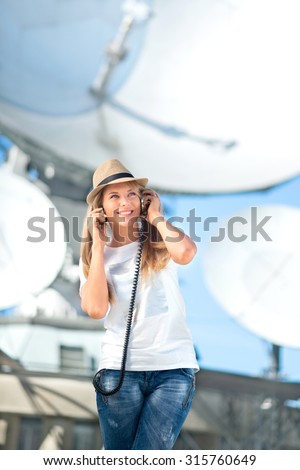 Happy young woman in hat listening to the music in vintage music headphones and dancing against background of satellite dishes that receive wireless signals from satellites.