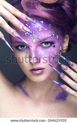 Girl with bright purple creative makeup with crystals and long nails. Beauty face. Picture taken in the studio on a gray background.