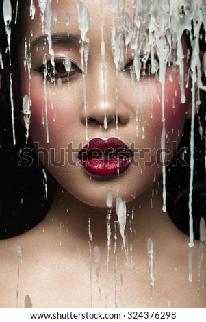 Beautiful Asian girl with bright make-up behind glass and drops of wax. Beauty face.