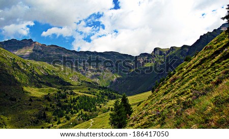 Mountain Panorama. A panoramic view of an hollow taken from an higher mountain side. There are pines and a road visible in the little valley below;  a cloudy blue sky on the background. Italian Alps.