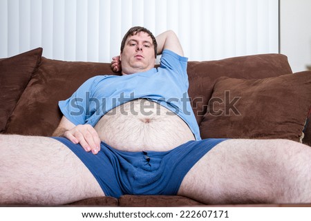 Young man sitting on couch with his legs open and his belly hanging out.