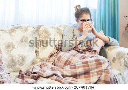 Sick young woman with headache sitting on the sofa and drinking tea. Girl with flu symptoms feeling tired. The flu and Ebola virus share similar symptoms, including headache, fever and fatigue.