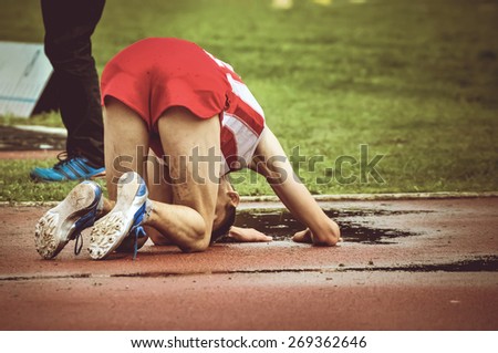 Exhausted young athlete falling on the ground after long run. Male runner on his knees on the running track after winning or loosing a race.
