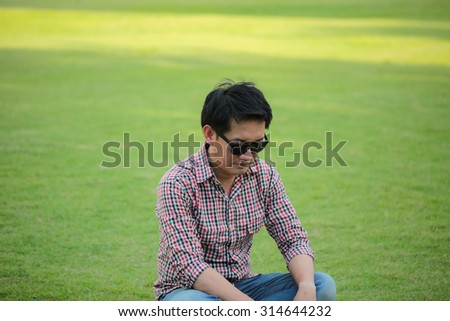 Man Wearing Check shirt, blue jeans sitting on green lawn wearing black sunglasses