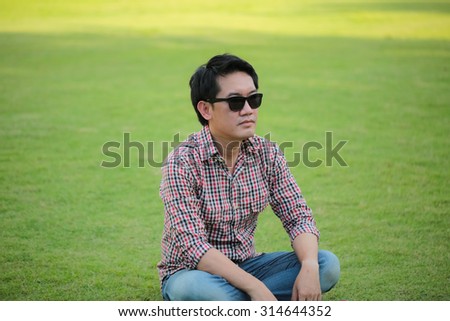 Man Wearing Check shirt, blue jeans sitting on green lawn wearing black sunglasses