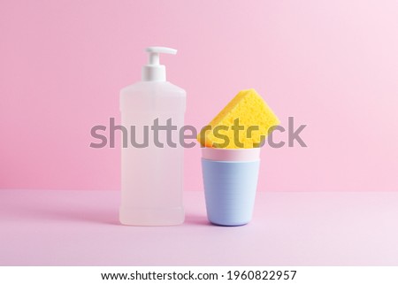 Creative still life with dishwashing supplies. White bottle of dishwash liquid and yellow kitchen cleaning sponge in stack of plastic cups. Front view, copy space, pink background.