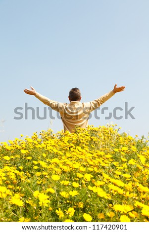 A happy young man is standing at a flowers field with his hands up.