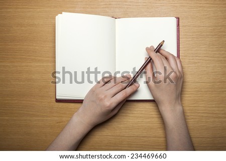 A female(woman) hands hold a blank(empty) book(note, diary) with pencil on the wood desk(table), top view at the studio.