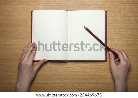 A female(woman) hands hold a blank(empty) book(note, diary) with pencil on the wood desk(table), top view at the studio.