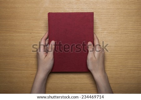 A female(woman) hands hold a blank(empty) red book(note, diary) cover on the wood desk(table), top view at the studio.