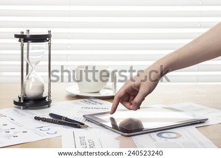 A working wooden desk(table) with tablet pc, coffee cup, graph, paper, sand timer, pencil and hand behind white blind(roller blind, shade).