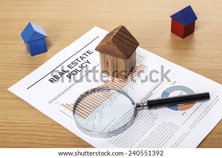 Three houses(made in wood block, blue and red) with a magnifier(reading glasses, magnifying glass[lens]) and graph paper(documents) on the office desk(table) behind white blind.