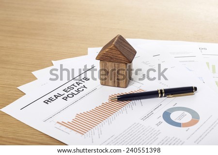 A house(made in wood block) with pen and graph paper(document) on the wood office desk(table) behind white blind.