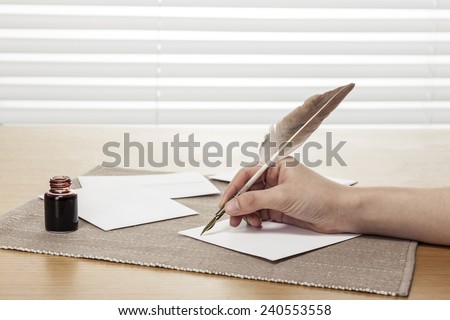 A female(woman) hand hold(write) a vintage feather quill pen with envelope and letter, ink on the office desk(table) behind white blind
