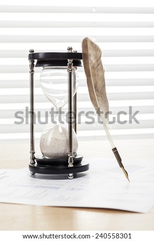 A sand timer(hour glass), feather quill pen, graph paper(document) on the wooden office desk(table) behind white blind.