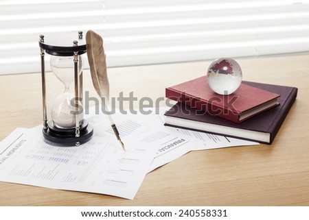 A sand timer(hour glass), feather quill pen, glass globe, graph paper(document) and book on the wooden office desk(table) behind white blind.
