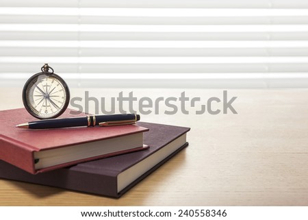 A vintage compass, pen, red book, diary, sketch book on the wooden office desk(table) behind white blind.