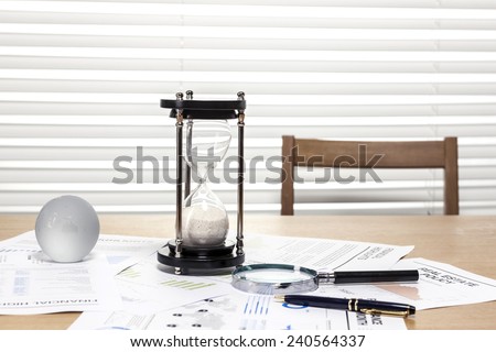 A sand timer(hour glass), glass globe, pen, magnifier, graph paper(document) on the wooden office desk(table) behind white blind.