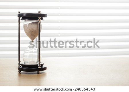 A sand timer(hour glass) on the wooden office desk(table) behind white blind.