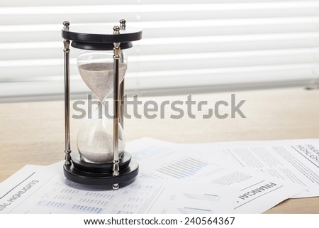 A sand timer(hour glass), graph paper(document) on the wooden office desk(table) behind white blind.
