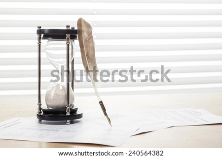 A sand timer(hour glass), feather quill pen, graph paper(document) on the wooden office desk(table) behind white blind.