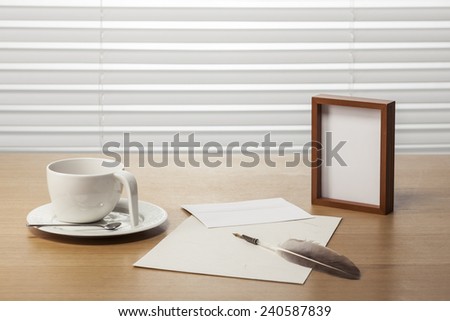 A coffee cup, feather quill pen, wood frame, letter, envelope on the office desk(table) behind white blind.