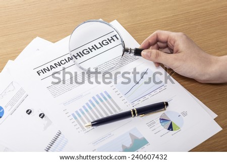 A female(woman) hand hold a magnifier(reading glass) point to graph paper(document), pen on the office desk(table).