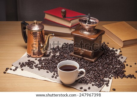 A brass coffee pot, grinder, white cup, beans and old(vintage, classic) books, table cloth on the wood table(desk).