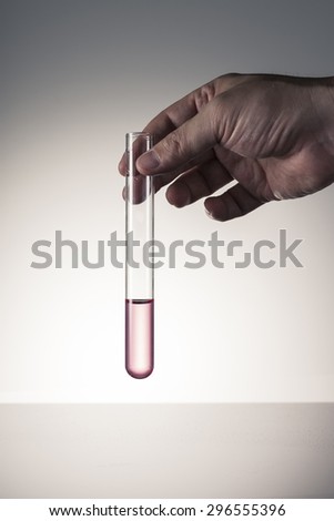 A man\'s hand hold a glass test tube(Proberohr) with red(magenta, pink) water(liquid, fluid) on the desk in the laboratory