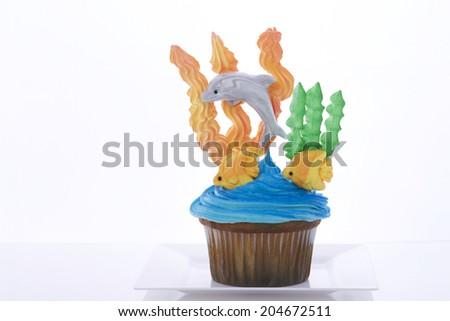 Aquatic under the sea themed cup cake created at home with royal icing coral and sea weed marshmallow fondant dolphins and fish. Presented on a white plate. Perfect for summer parties