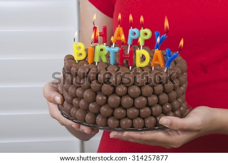 Young girl holding Chocolate Birthday Cake decorated with candy malt balls in front of her with happy birthday candles burning. Fast and easy home made cake for children or adult birthday party